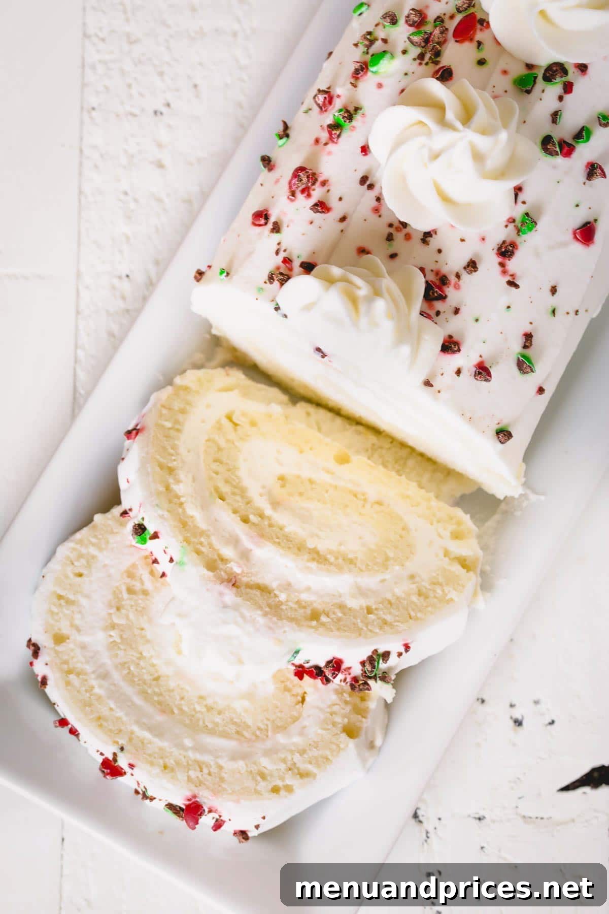 Enchanted Apple Cream Roll 8 A beautifully sliced white roll cake, revealing its smooth, creamy mousse filling and delicate sponge, adorned with crushed red and green M&Ms on top.