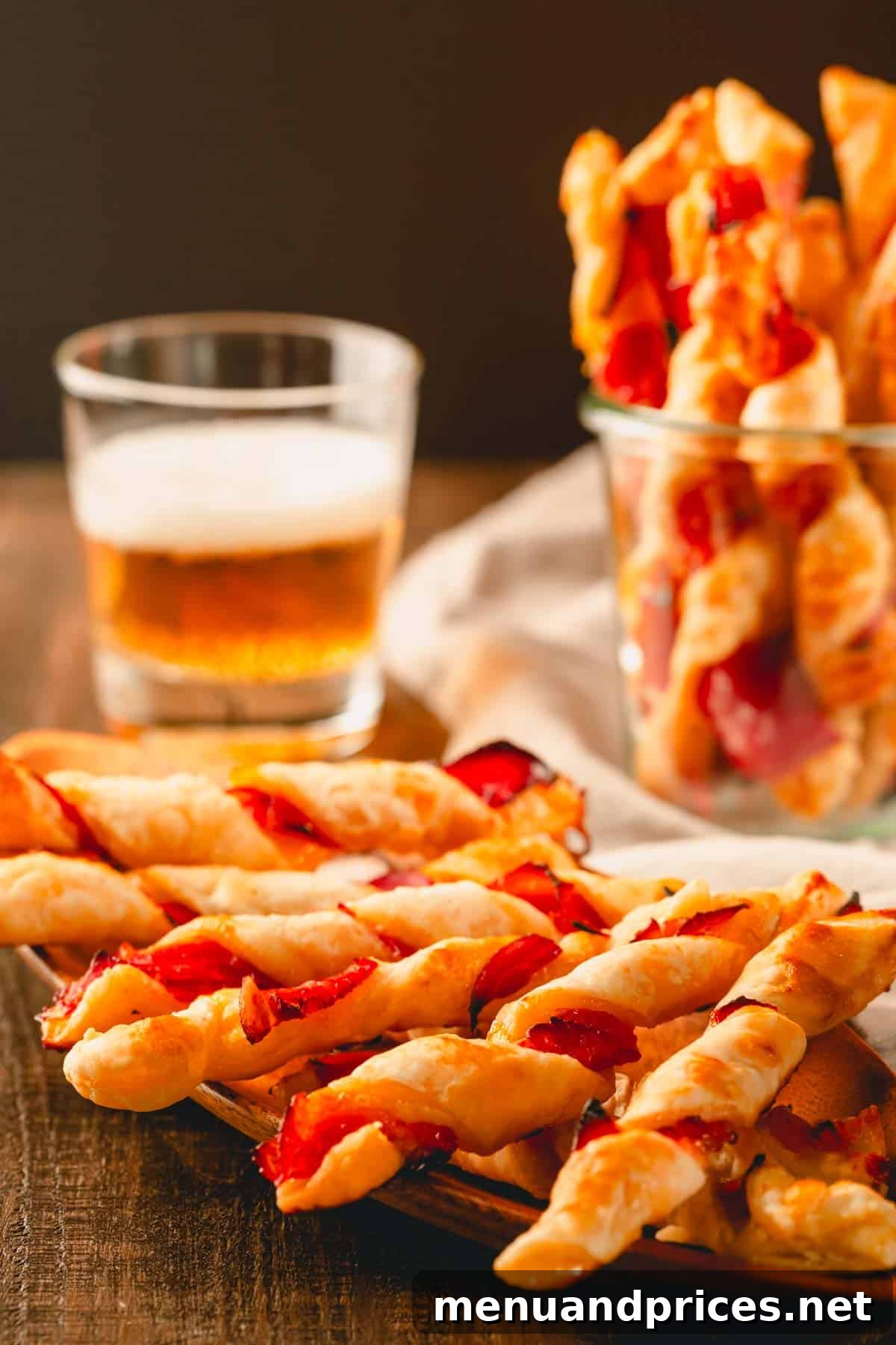 Golden Savory Spirals 12 Prosciutto puff pastry twists on a plate and in a glass cup with a glass of beer in the background.