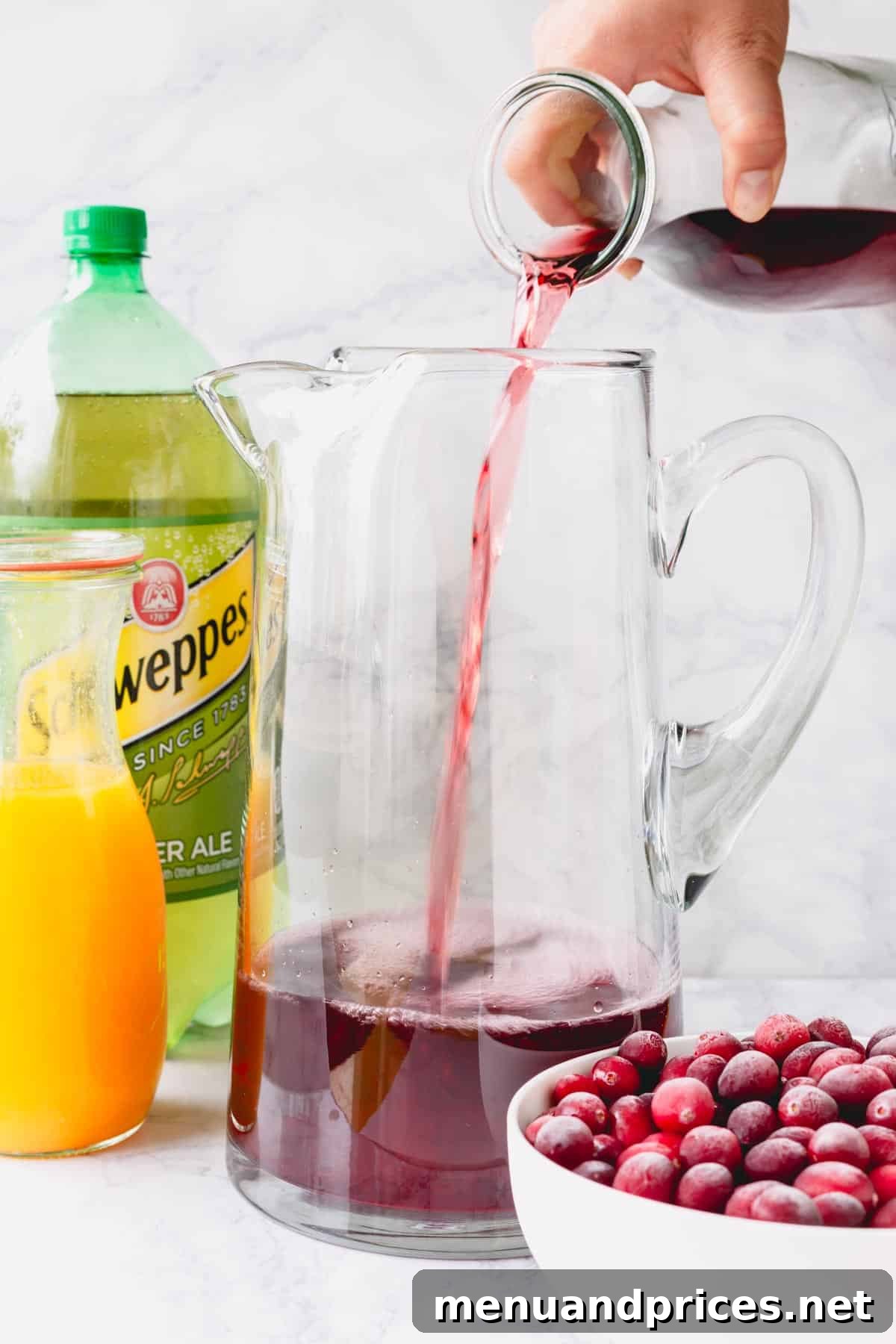 Cranberry juice being poured into a pitcher with ginger ale, orange juice, and pomegranates next to it, illustrating the first step of mixing.