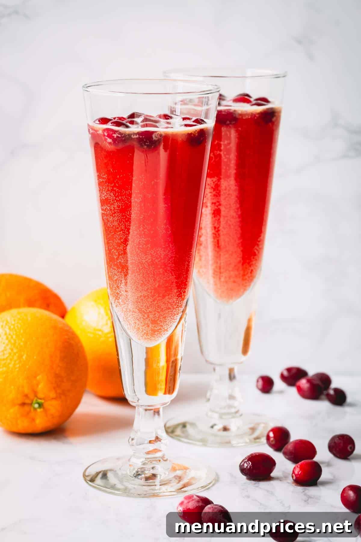 Two glasses of orange cranberry spritzer with oranges and pomegranates scattered around, creating a festive scene.