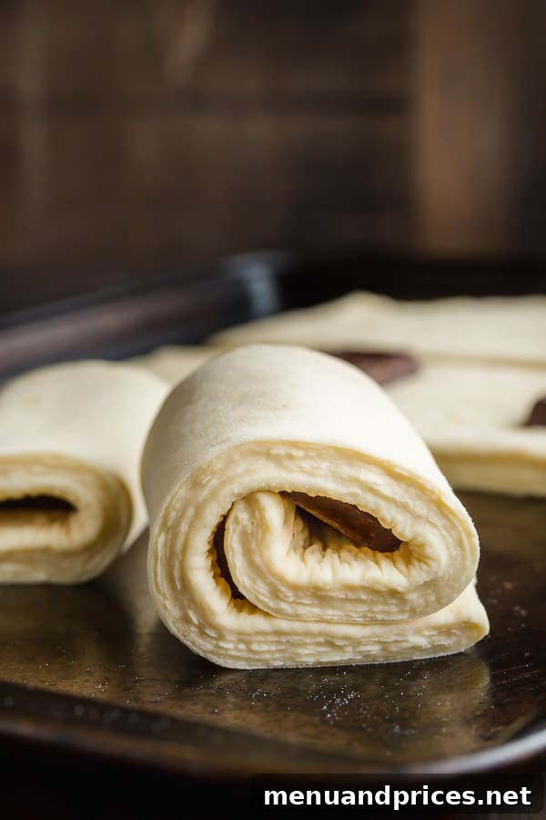 Unbaked croissant dough carefully wrapped around two pieces of chocolate batons, positioned on a baking sheet, patiently awaiting the final proofing stage before its journey into the oven.