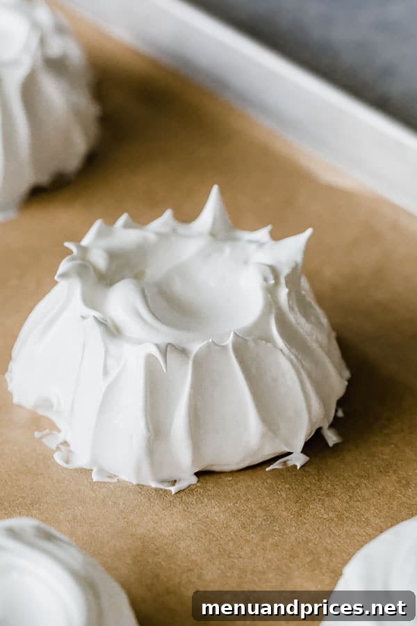 Two styles of mini pavlova meringue nests on a baking sheet, one with swiped edges, the other piped.