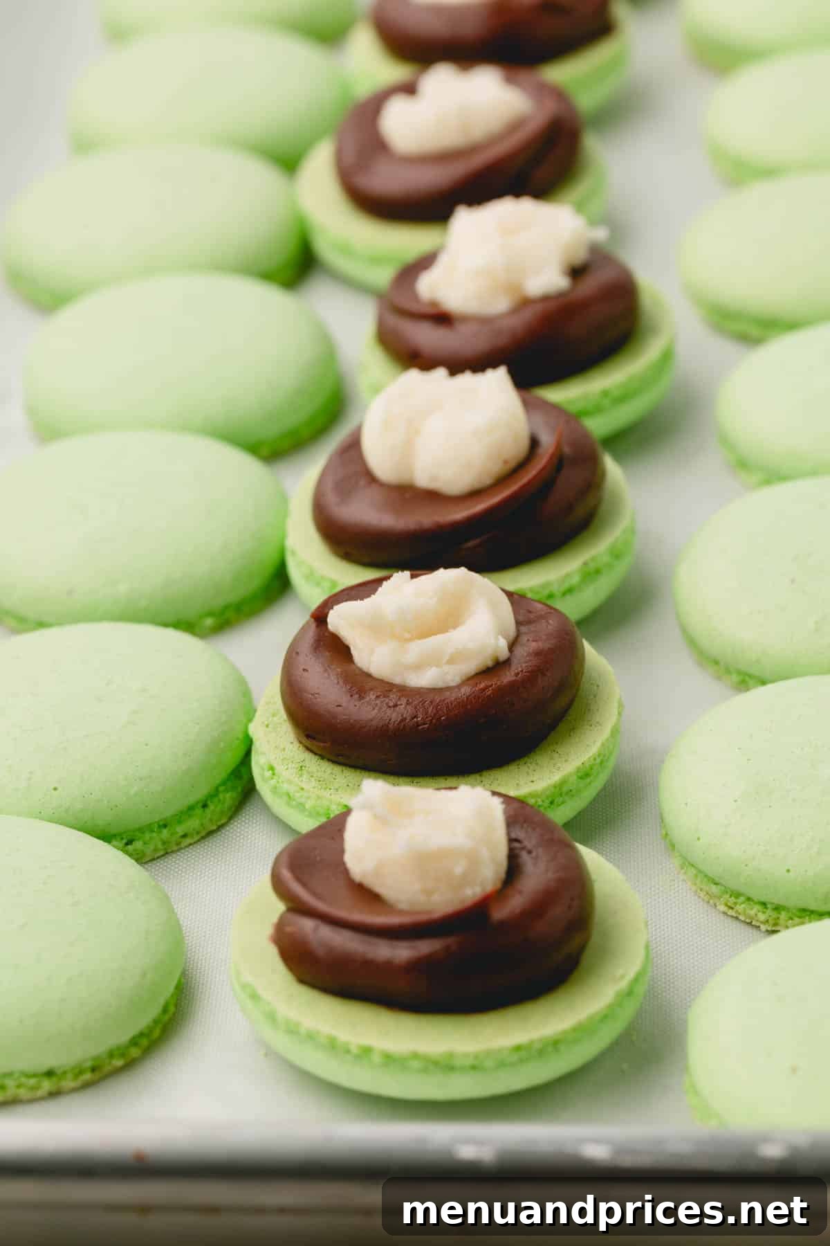 Minty Cocoa Macaron Bliss 21 Green macaron shells, each with a perfect ring of chocolate ganache and a spoonful of creamy white mint candy filling precisely placed in the center, ready for their top shell.