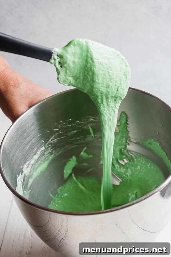 Minty Cocoa Macaron Bliss 10 Smooth green macaron batter, showing the ideal 'lava-like' consistency, slowly drips from a silicone spatula into a stainless steel mixing bowl.
