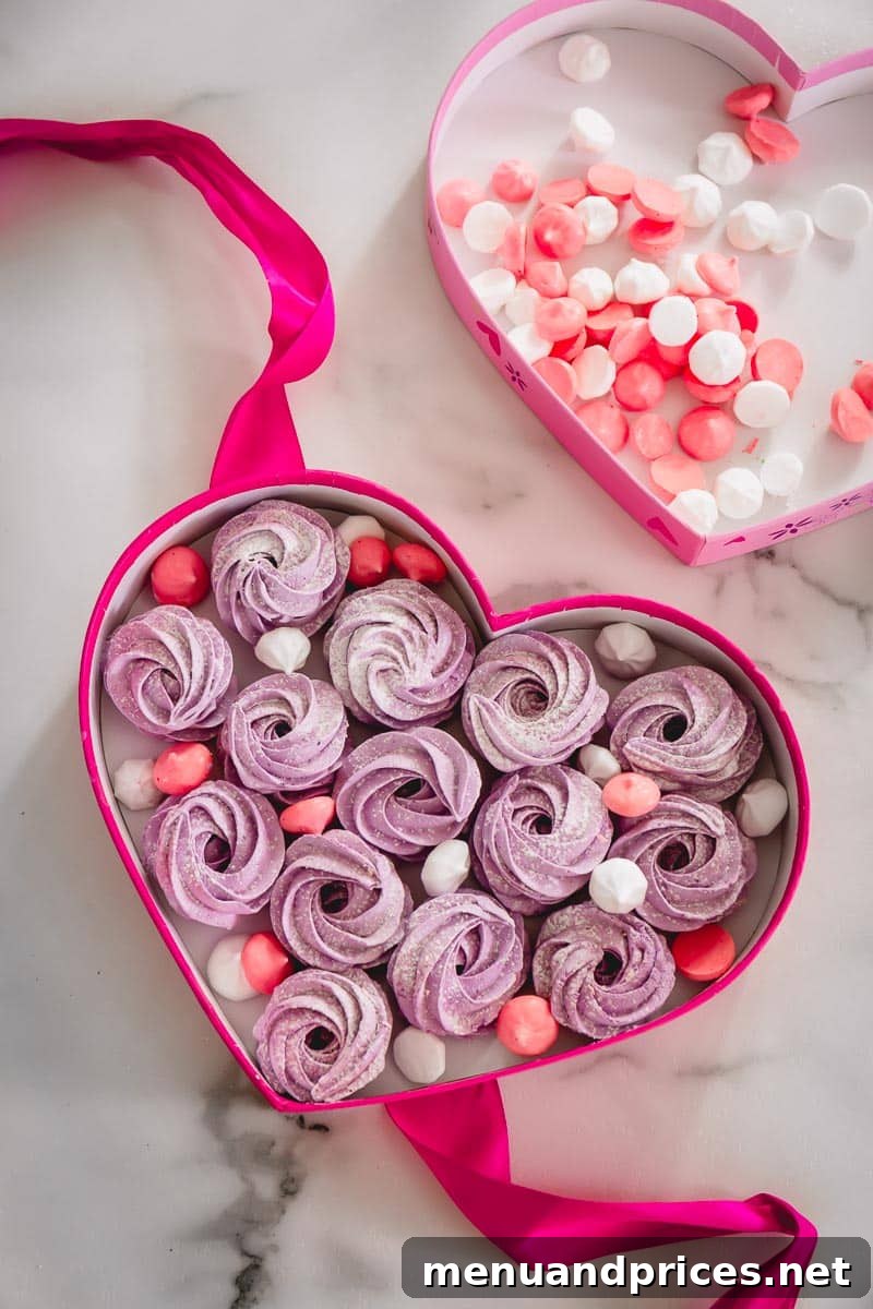 A charming pink heart-shaped box filled with an assortment of vibrant purple marshmallow rosettes, alongside delicate mini red and pink meringue kiss cookies, creating a delightful confectionery display.