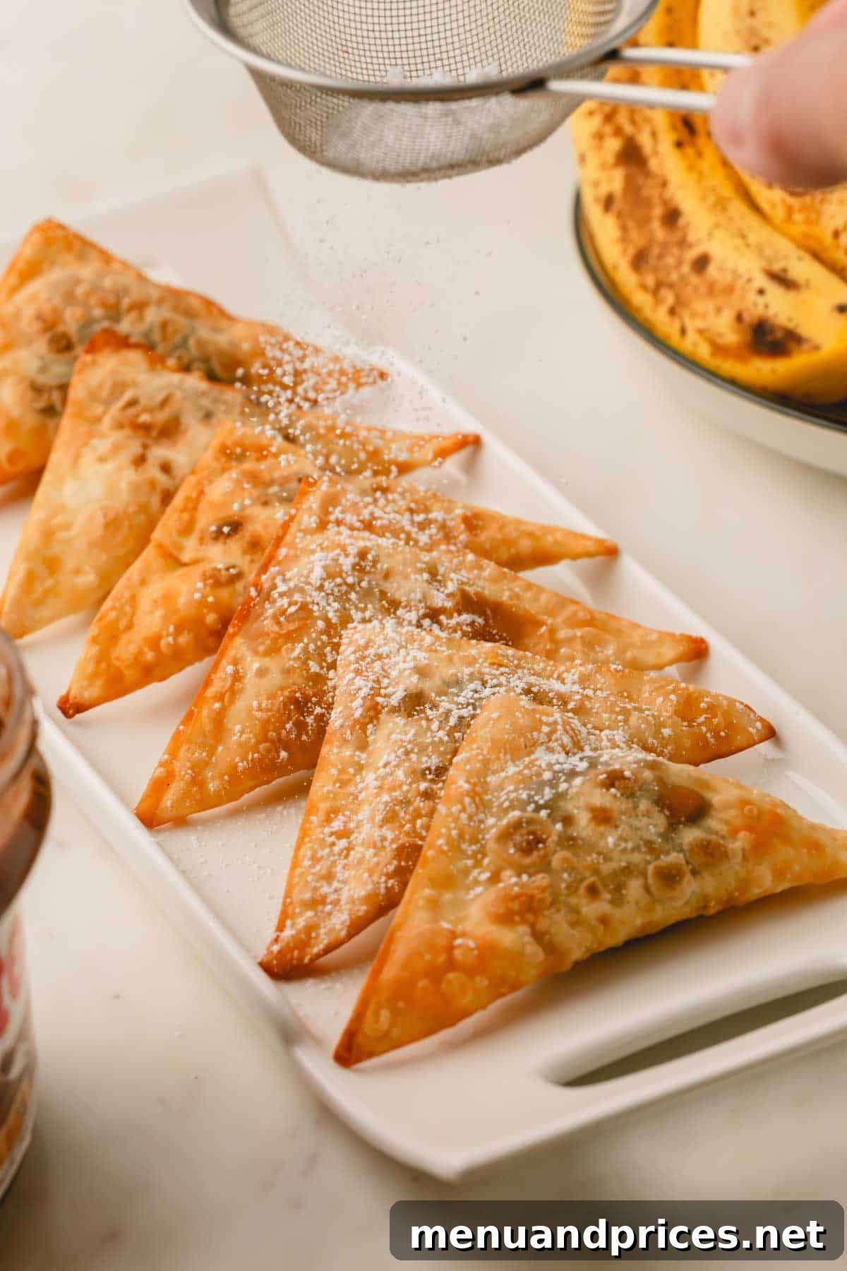 A beautiful platter of golden-brown banana and Nutella wontons, freshly baked and being dusted generously with powdered sugar.