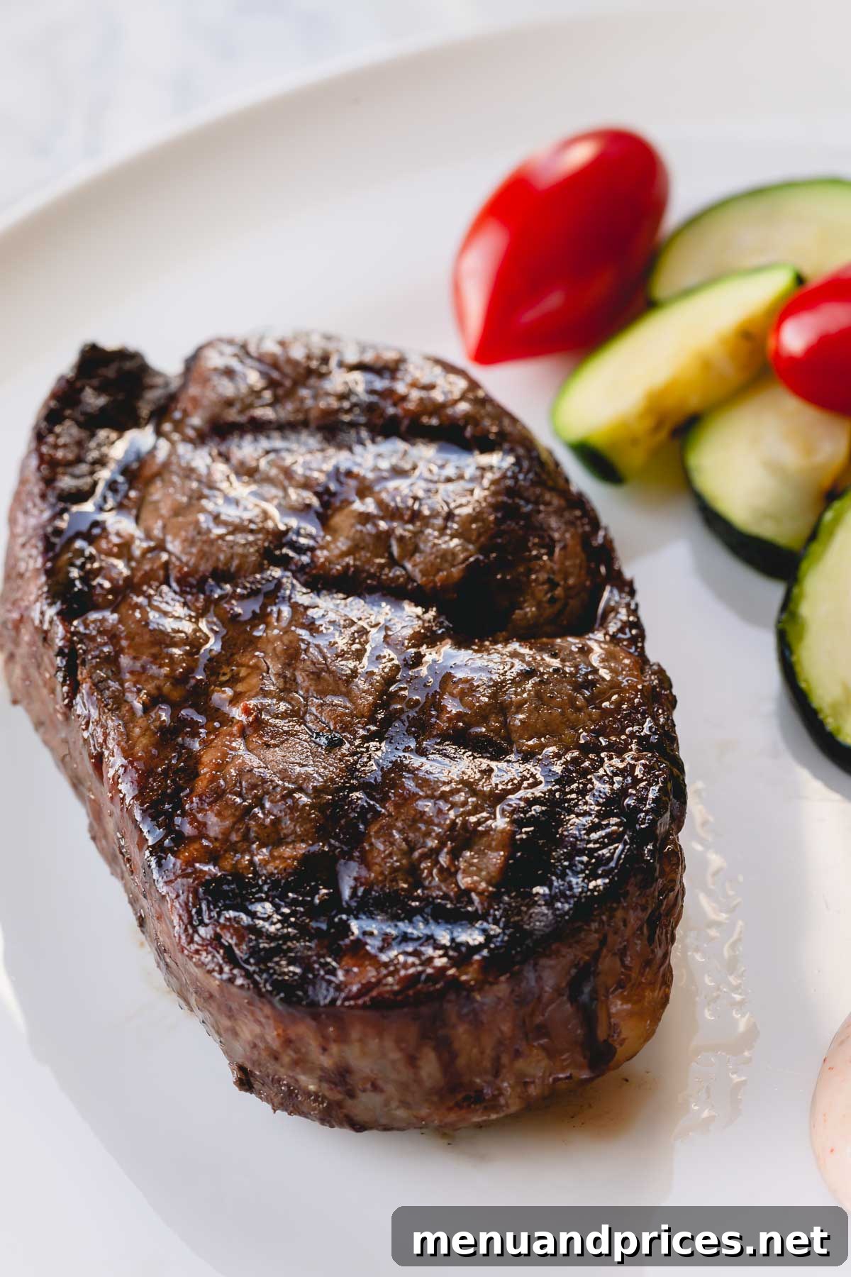 Effortless Grilled Filet Mignon 3 A whole grilled filet mignon on a white plate with zucchini and cherry tomatoes on the side.