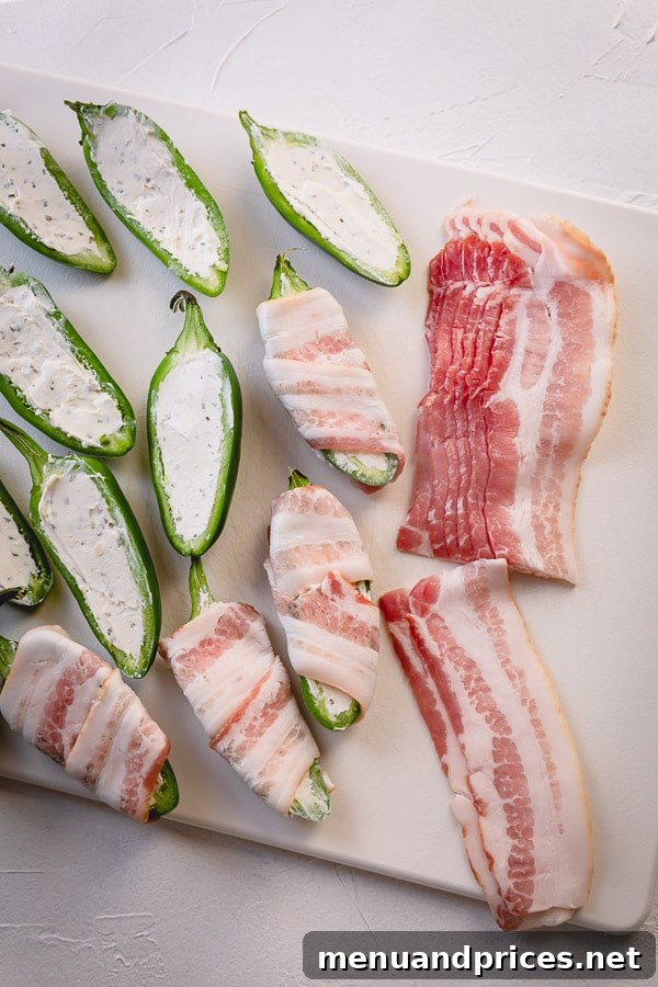 Bacon strips being wrapped around the cream cheese-filled jalapeno halves.