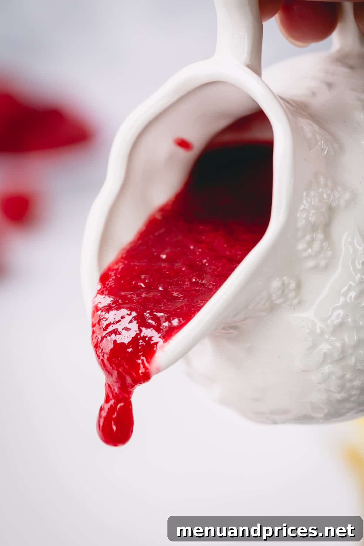 Velvety Raspberry Sauce Without the Seeds 3 Smooth raspberry sauce being elegantly poured from a white sauce pitcher into a pristine white serving bowl, highlighting its perfect, pourable consistency.