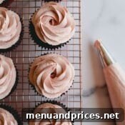 Chocolate cupcakes on a wire rack topped with strawberry whipped cream frosting.