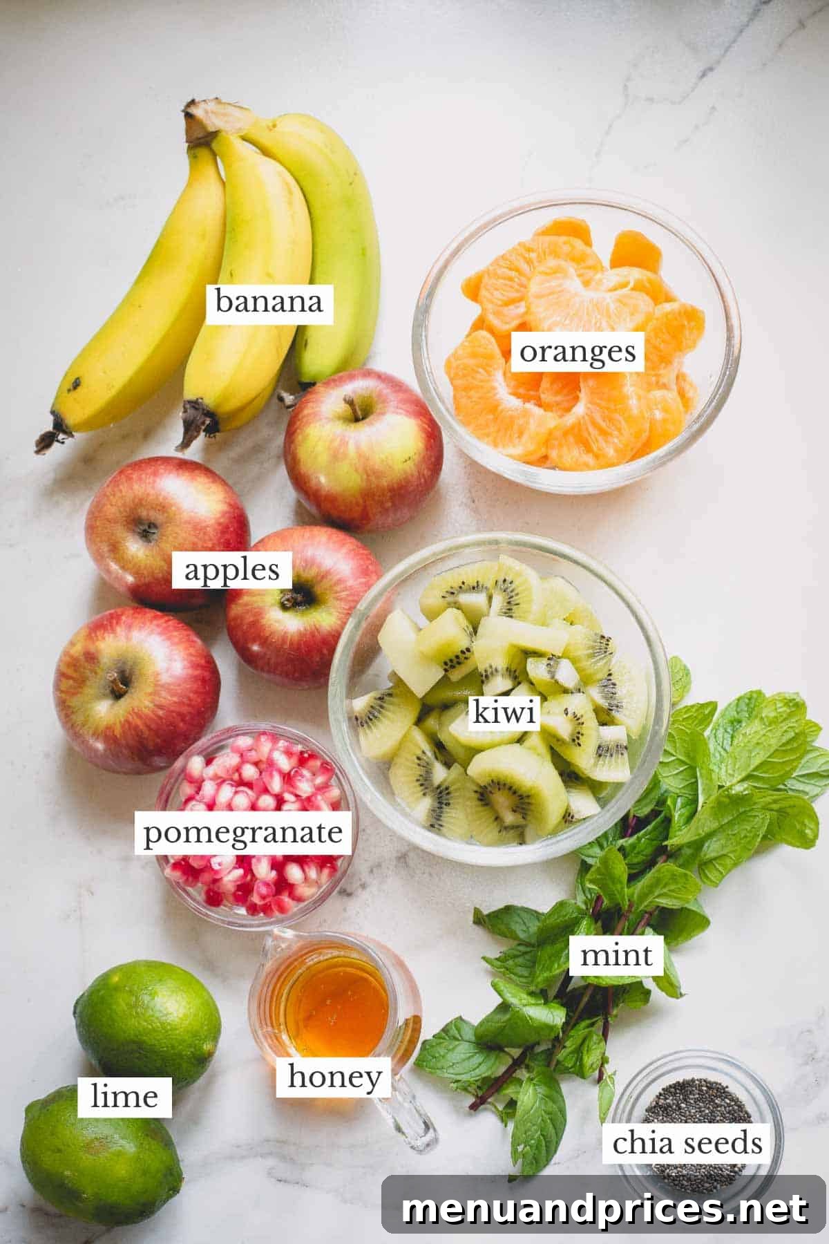 A flat lay photograph of fresh ingredients for fruit salad: bananas, apples, mandarin oranges, kiwi, fresh mint leaves, whole limes, and a jar of honey.