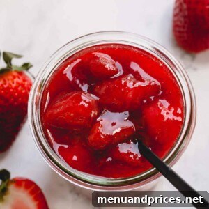 Overhead image of a spoon sticking out of a glass jar full of chunky strawberry sauce.