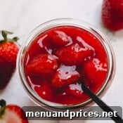 Overhead image of a spoon sticking out of a glass jar full of chunky strawberry sauce.