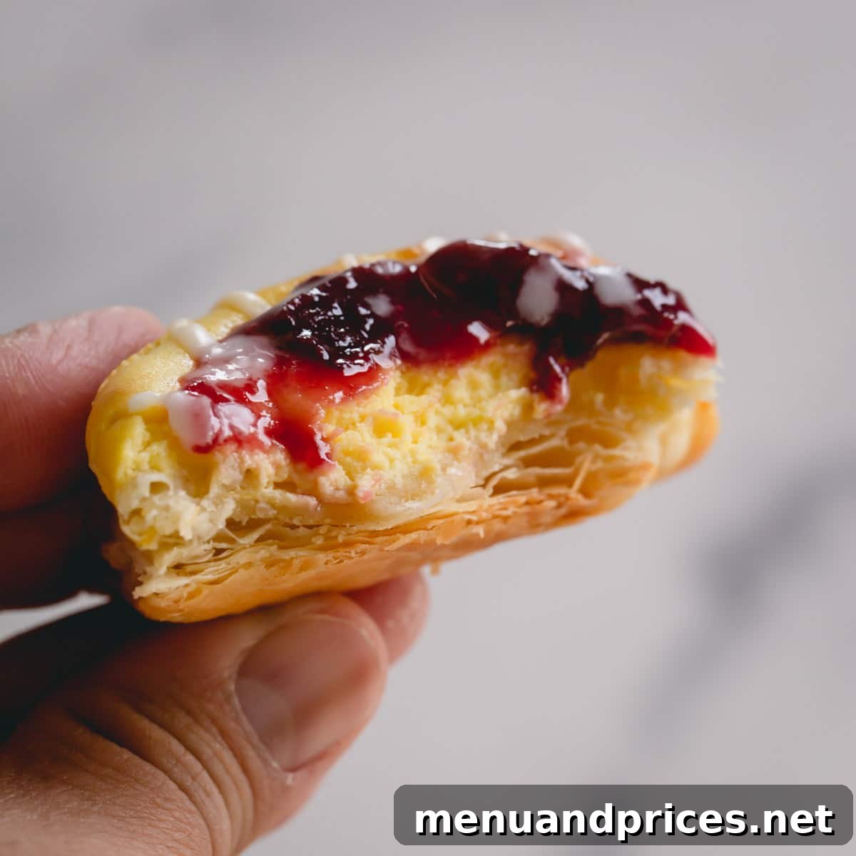 A hand holding a freshly baked cherry danish with a bite taken, revealing the creamy cheese center, flaky pastry layers, and sweet cherry filling.