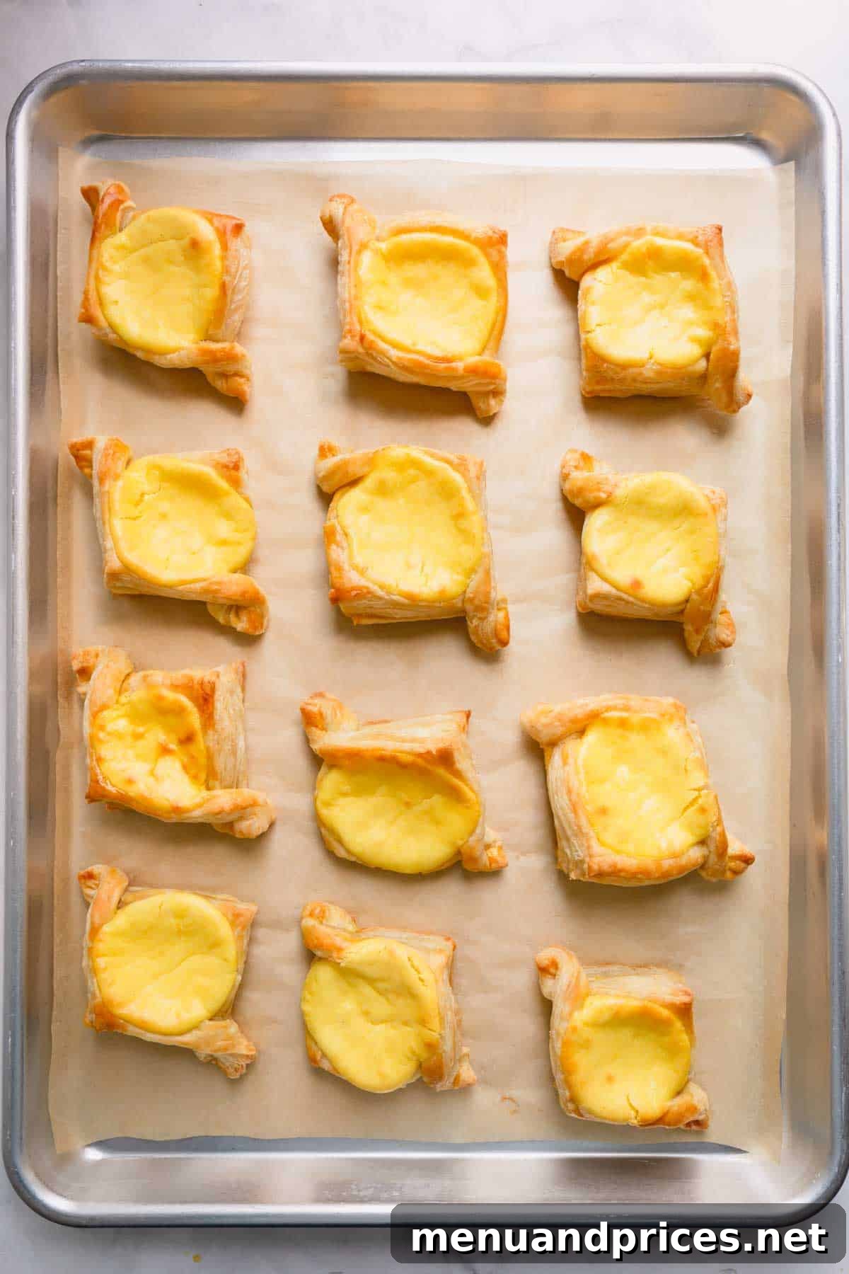 Freshly baked puff pastry danishes, golden and perfectly puffed, with a creamy cheese filling visible, cooling on a parchment paper-lined baking sheet.