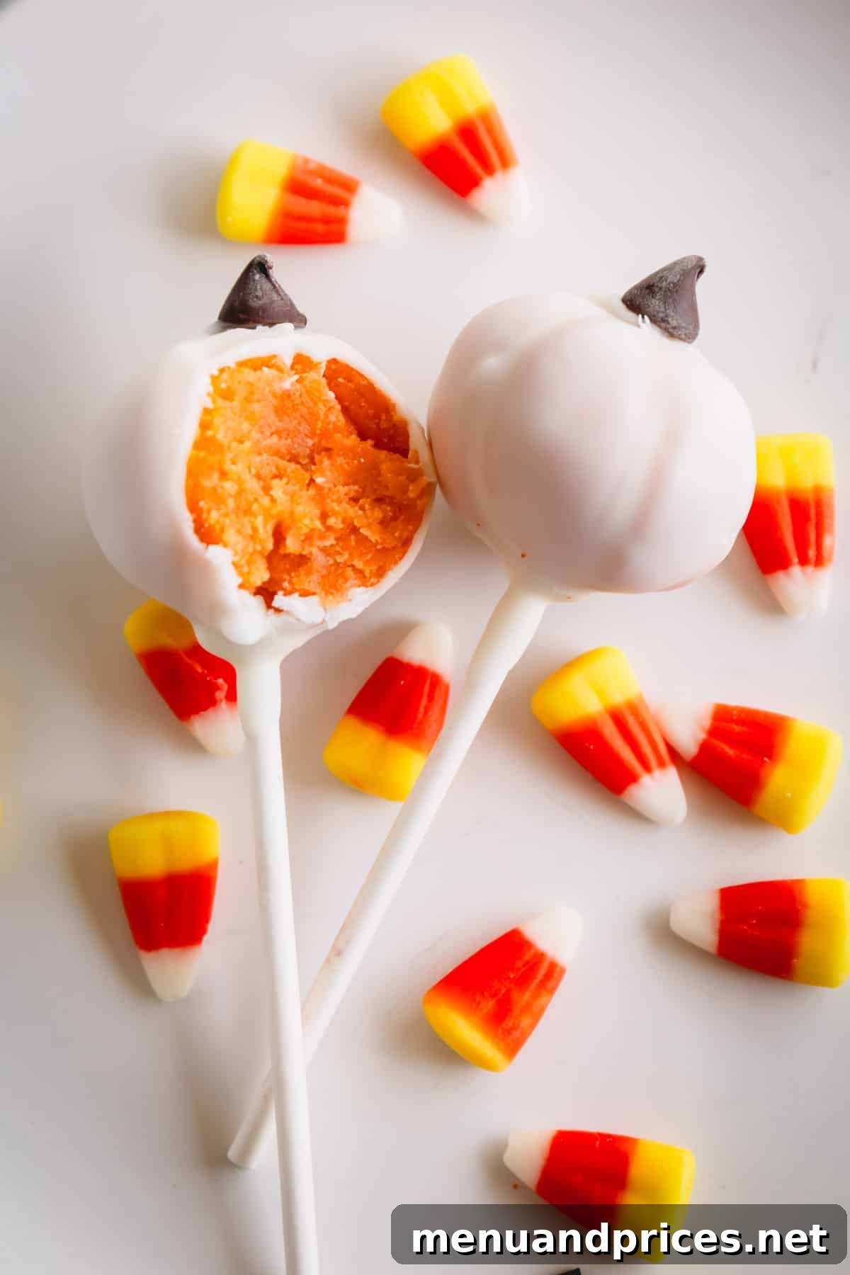 Two delightful white pumpkin cake pops, with one showing a bite taken out, surrounded by festive candy corn.
