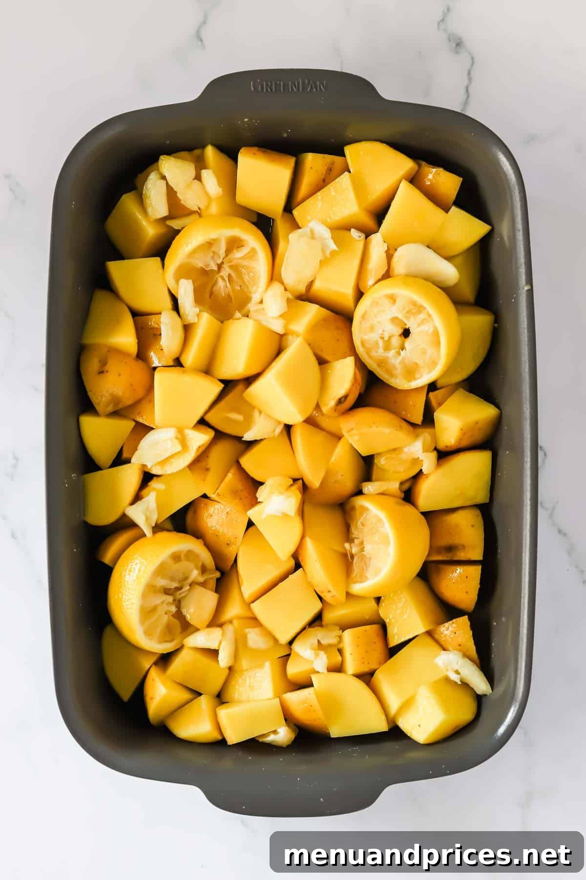A baking dish filled with neatly arranged chopped potatoes, whole smashed garlic cloves, and bright lemon halves, all seasoned and ready for the chicken.