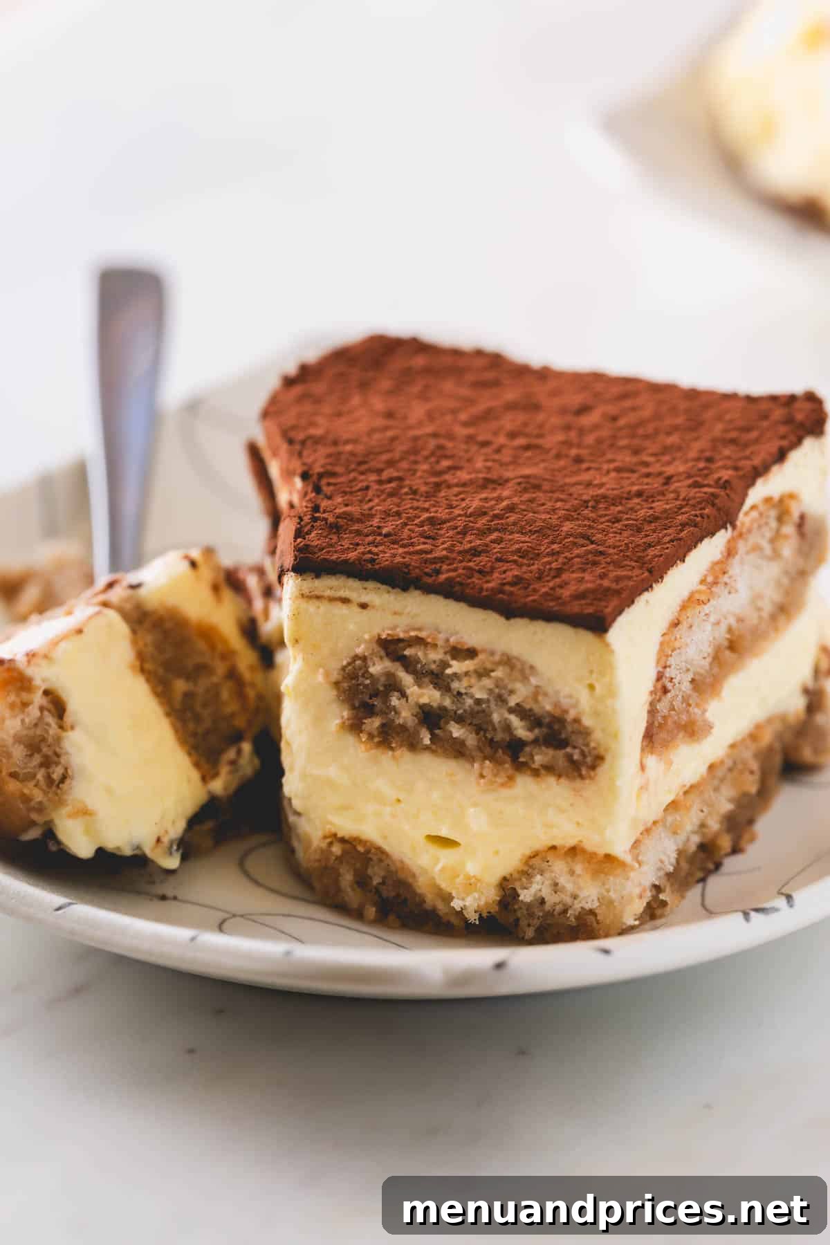 Authentic Italian Tiramisu 2 A perfectly portioned slice of classic tiramisu sits elegantly on a white plate, with a fork poised to capture the first delicious bite. The distinct layers of coffee-soaked ladyfingers and creamy mascarpone are clearly visible, topped with a dusting of cocoa.