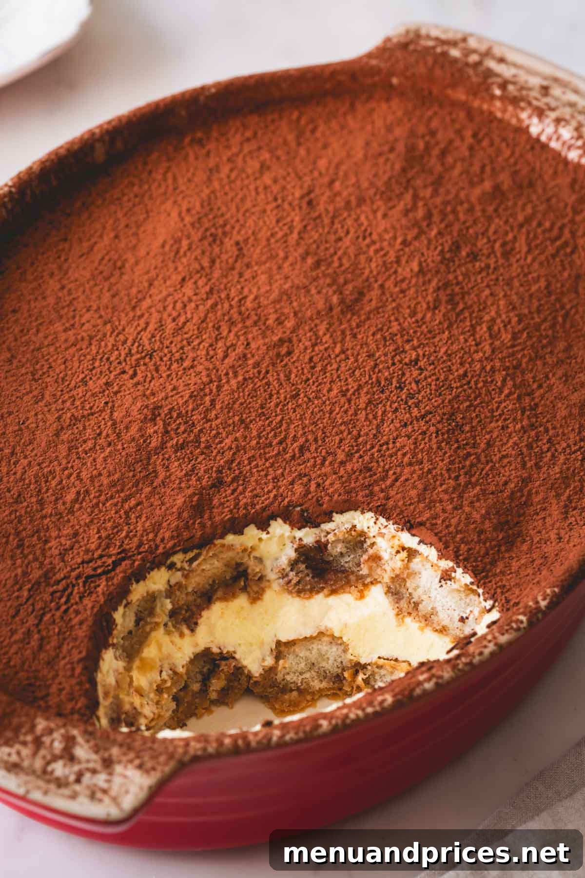 Authentic Italian Tiramisu 3 A ceramic dish filled with freshly made tiramisu, with one generous serving already scooped out, revealing the delightful layers within.