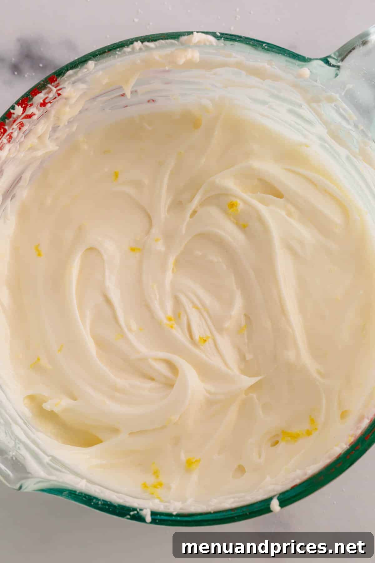 A measuring cup filled with vibrant lemon cream cheese frosting, garnished with delicate lemon zest flakes.