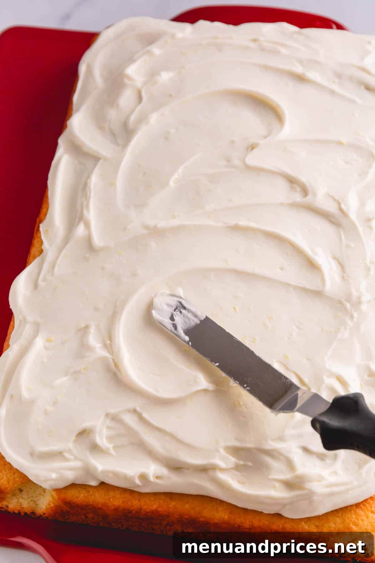Sunshine Lemon Sheet Cake 11 A beautiful lemon sheet cake in a baking pan, being expertly frosted with a generous layer of creamy lemon cream cheese frosting using an offset spatula, creating a smooth and inviting finish.