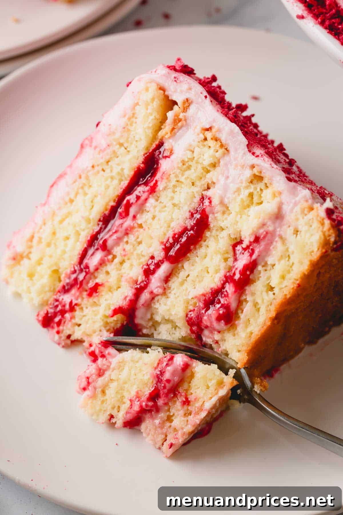 A fork taking a bite from a slice of raspberry layer cake on a plate, highlighting its texture.