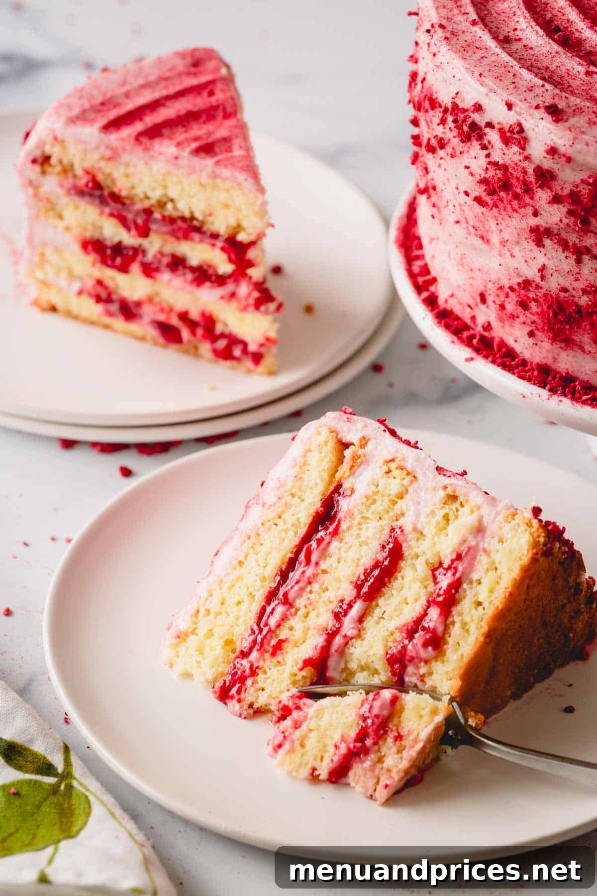 Two slices of raspberry layer cake on plates, next to the whole cake, with a fork cutting a bite from one slice.