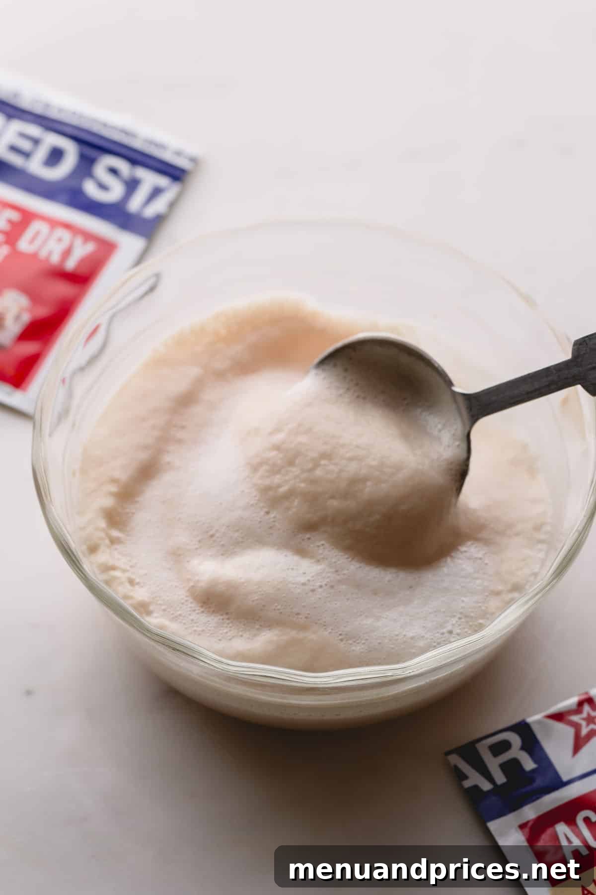 Yeast Essentials: Types and Their Ideal Uses 9 A close-up view of a small glass bowl showcasing a vibrant, foamy yeast mixture, with a spoon gently dipped into it, and a packet of active dry yeast partially visible in the background, confirming successful activation.