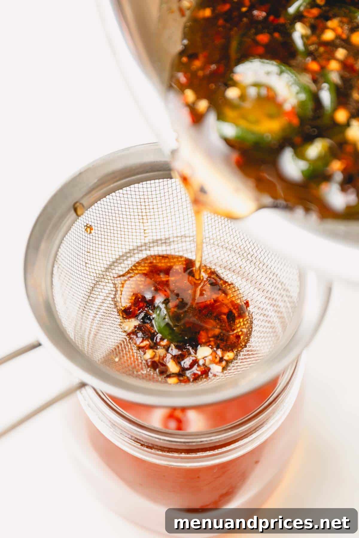 Sweet Heat Honey Formula 6 Freshly made hot honey being strained through a fine-mesh sieve into a glass jar, removing the solids for a smooth consistency.