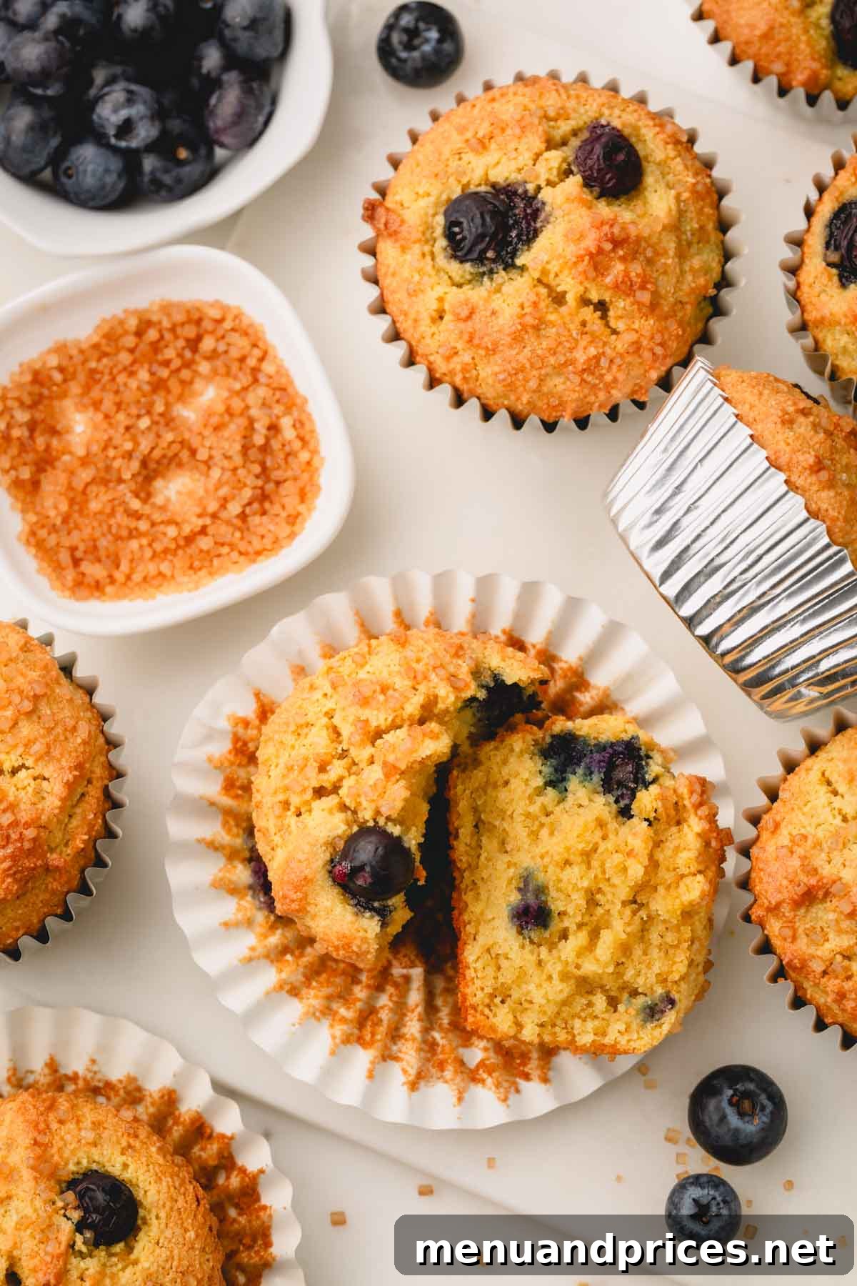Moist Almond Flour Blueberry Muffins 3 An almond flour blueberry muffin split in half, revealing its moist interior and numerous blueberries, placed on a muffin liner surrounded by other whole muffins.