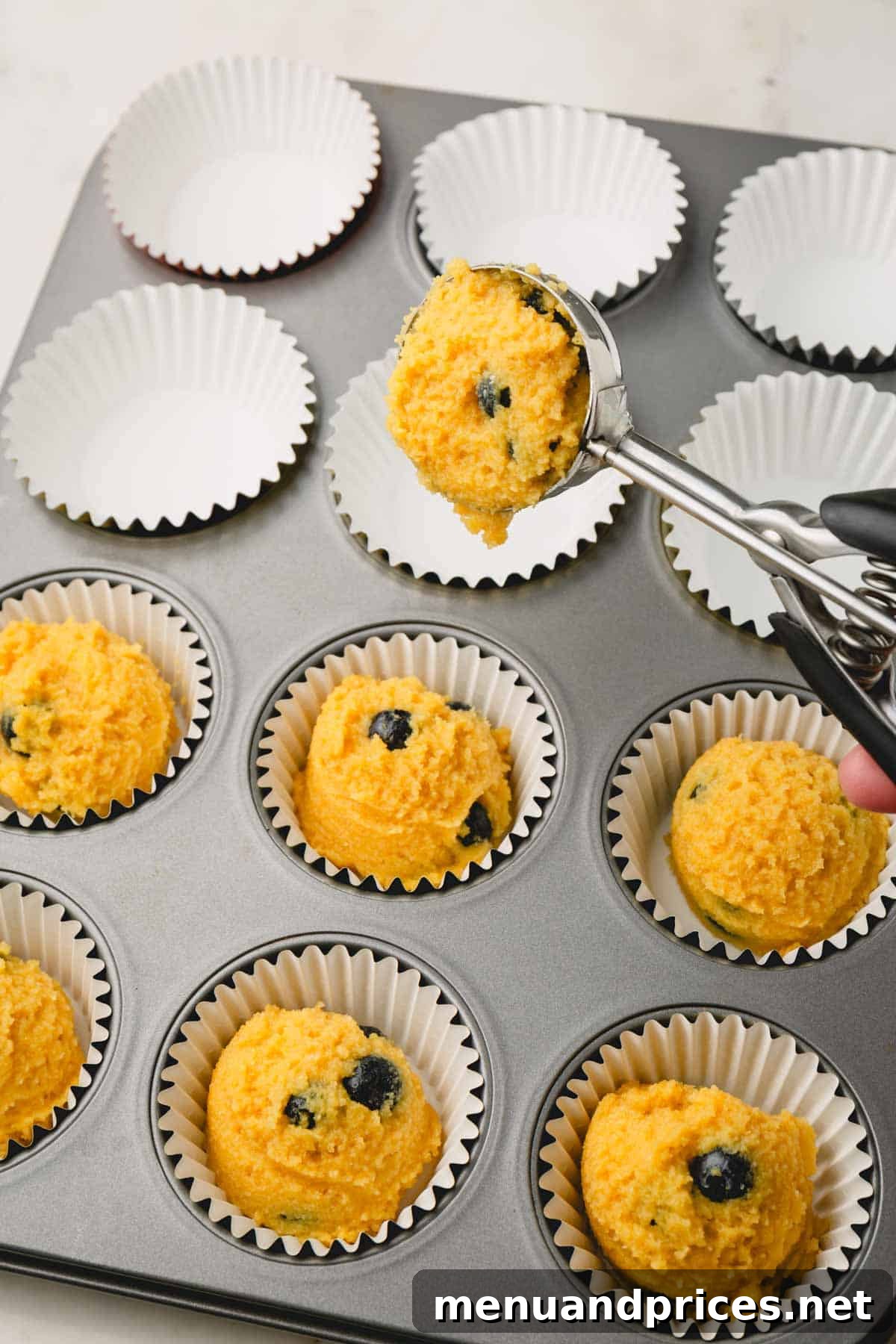 Moist Almond Flour Blueberry Muffins 10 Almond flour blueberry muffin batter being carefully portioned into a muffin tin lined with paper muffin liners, filled almost to the brim.