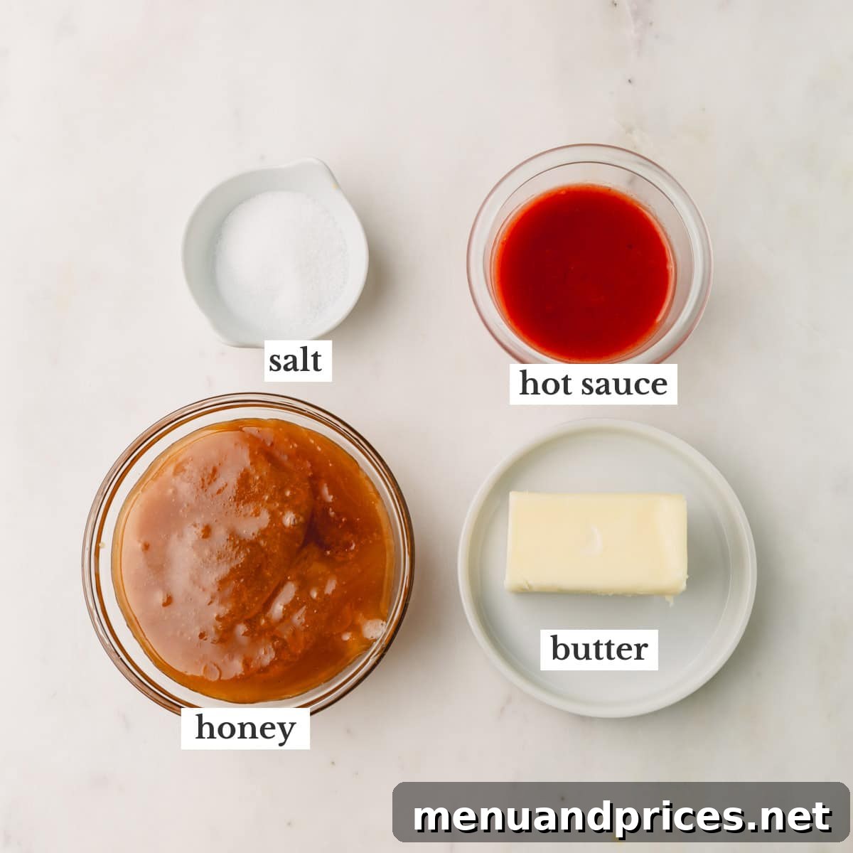 Golden Blaze Drizzle 3 A flat lay photograph showcasing the four key ingredients for hot honey sauce: a stick of butter, a bottle of hot sauce, a jar of honey, and a small pile of coarse kosher salt.