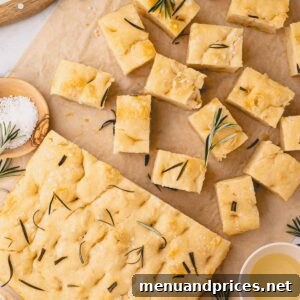 Foolproof Homemade Focaccia 18 Half a loaf of rosemary focaccia bread and the rest of the bread sliced into squares on parchment paper, showcasing its crisp crust and soft interior.
