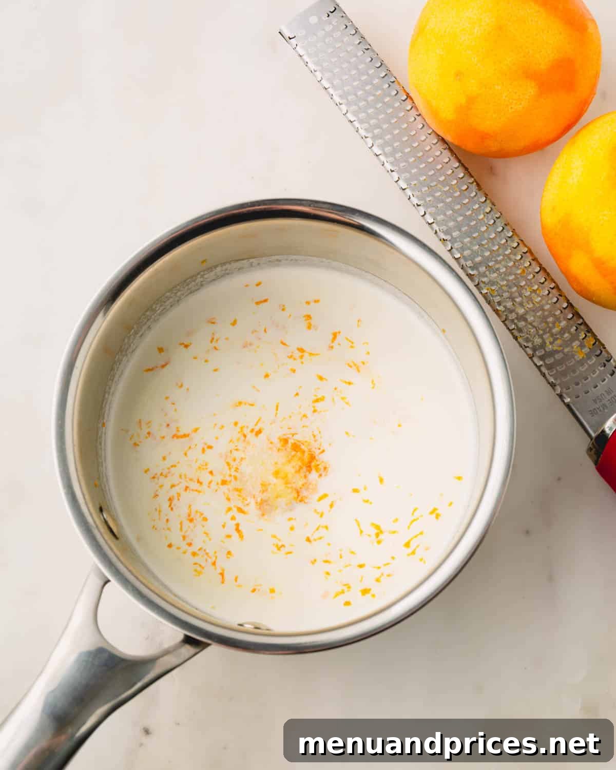 A pot of gently warmed cream infused with fresh orange zest, next to a zester and two whole oranges.