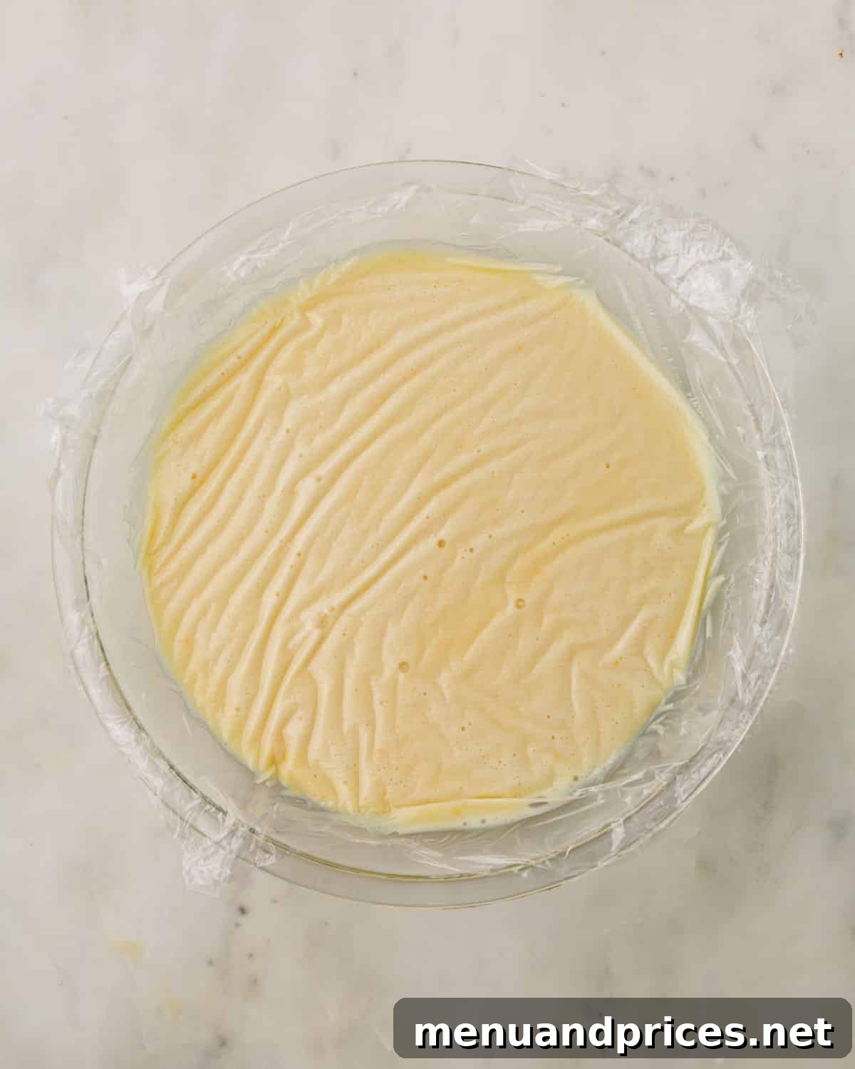 A bowl of white chocolate orange ganache covered with plastic wrap, ensuring it touches the surface.
