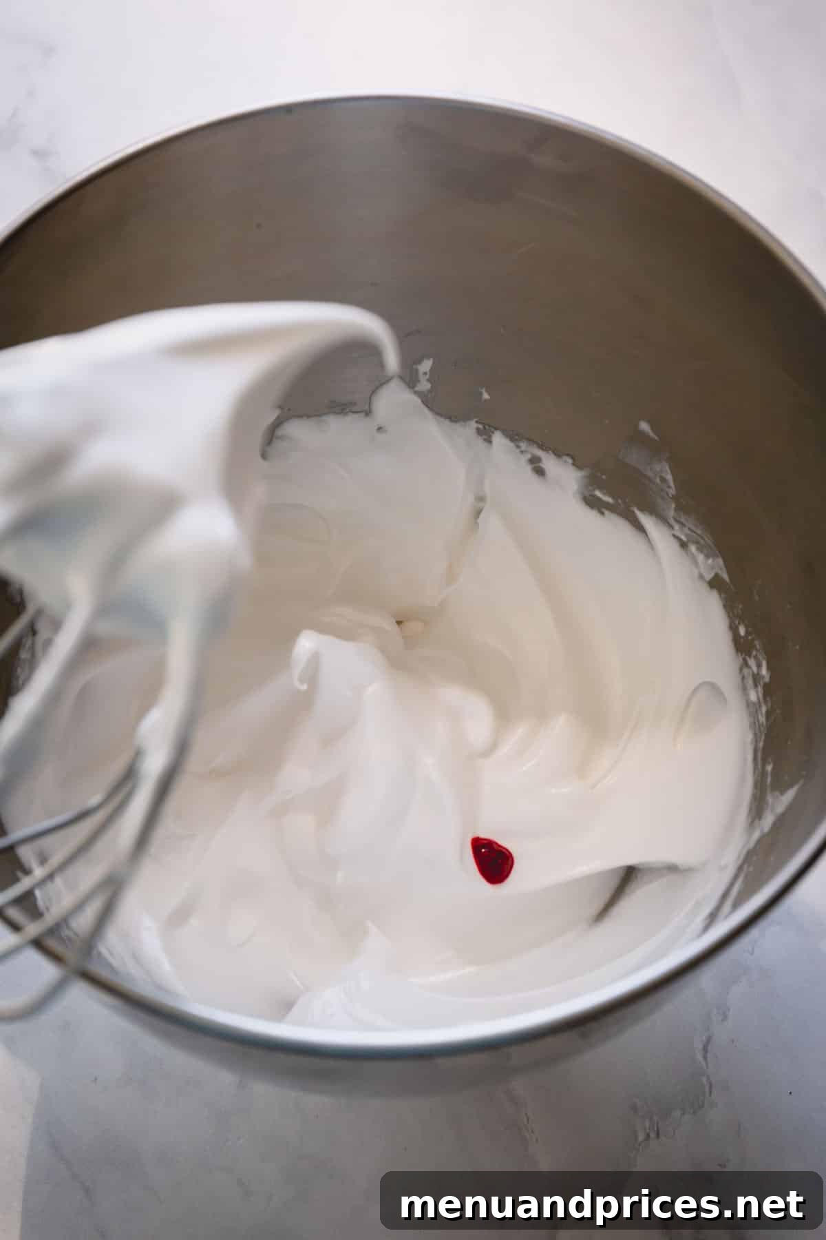 Meringue at the soft peak stage in a mixer bowl, with a whisk attachment and a few drops of vibrant burgundy food coloring.