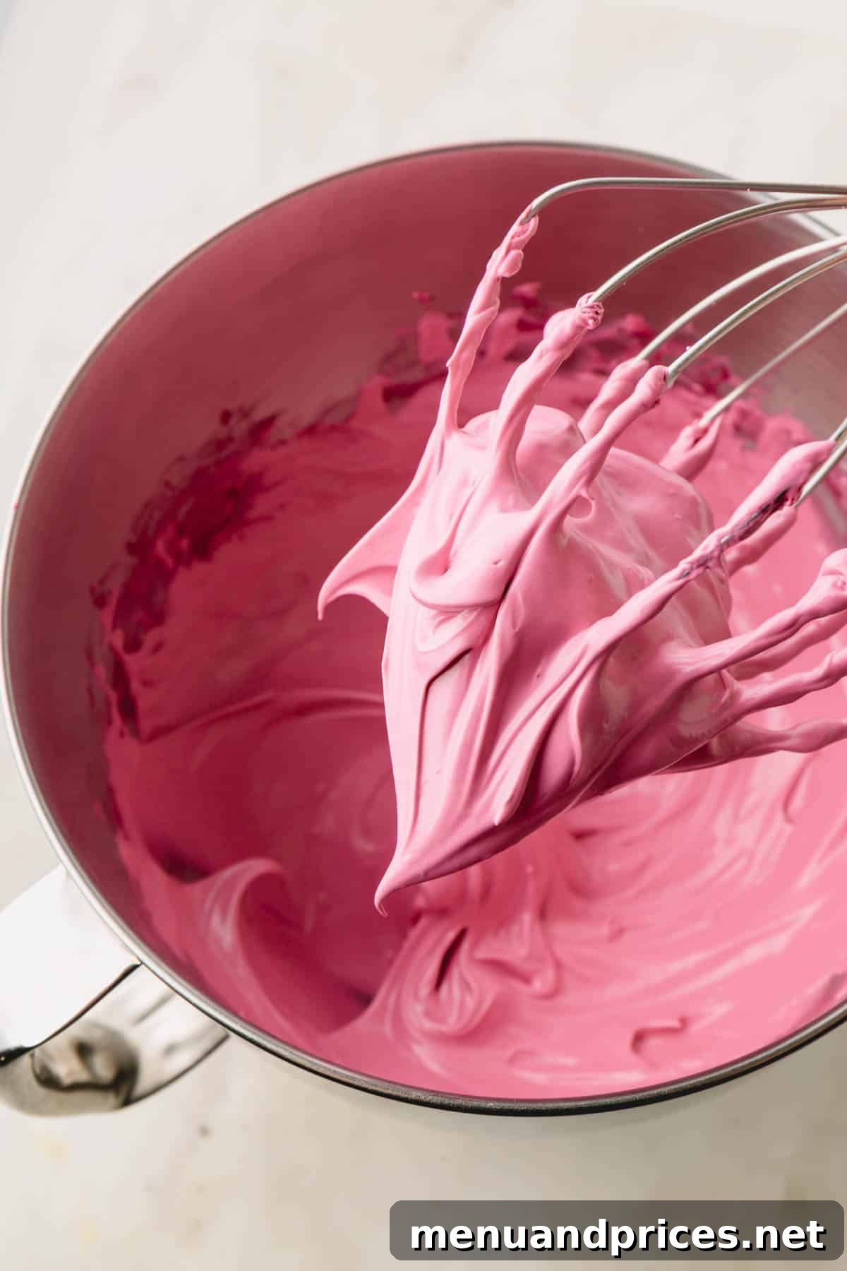A bowl filled with beautiful pink meringue, with a whisk attachment holding stiff peaks suspended above it.