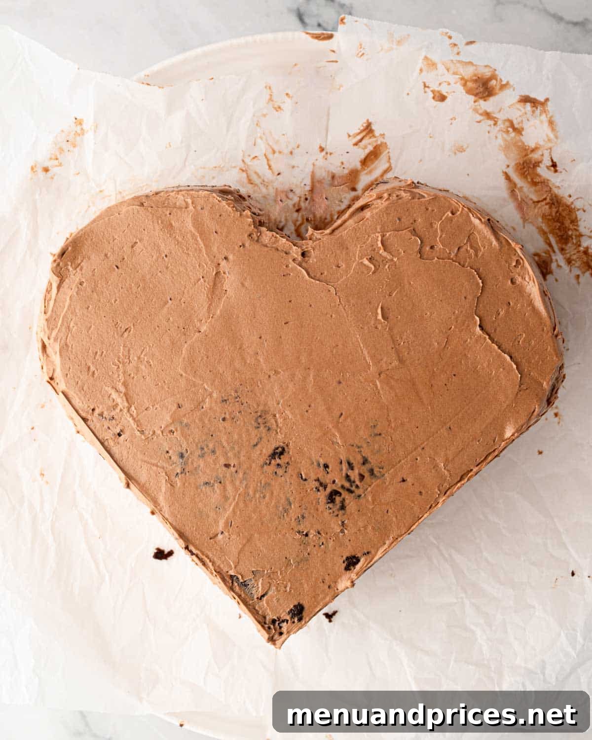 A chocolate heart cake being covered with a thin crumb coat of chocolate Swiss meringue buttercream.