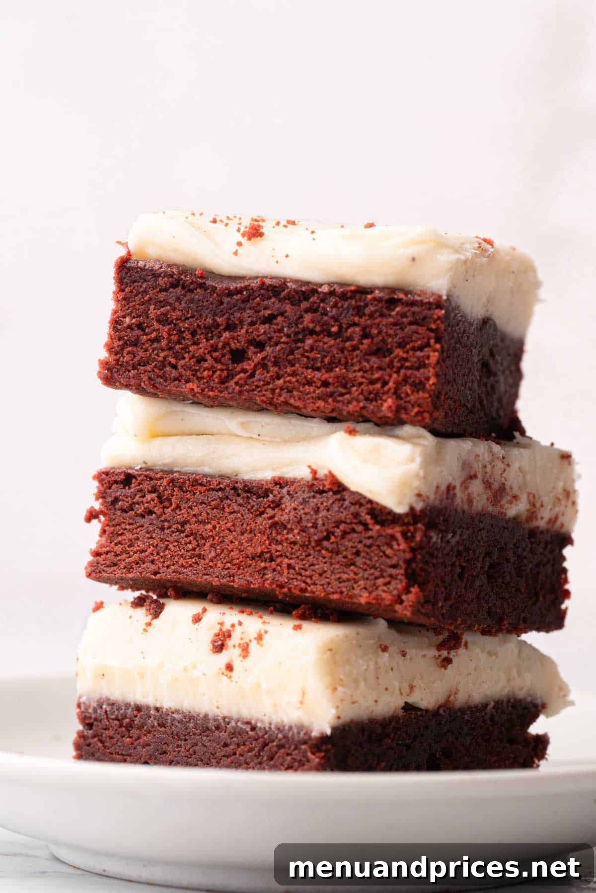 Crimson Velvet Brownies 2 A stack of three red velvet brownies with cream cheese frosting on a plate, ready to be enjoyed.