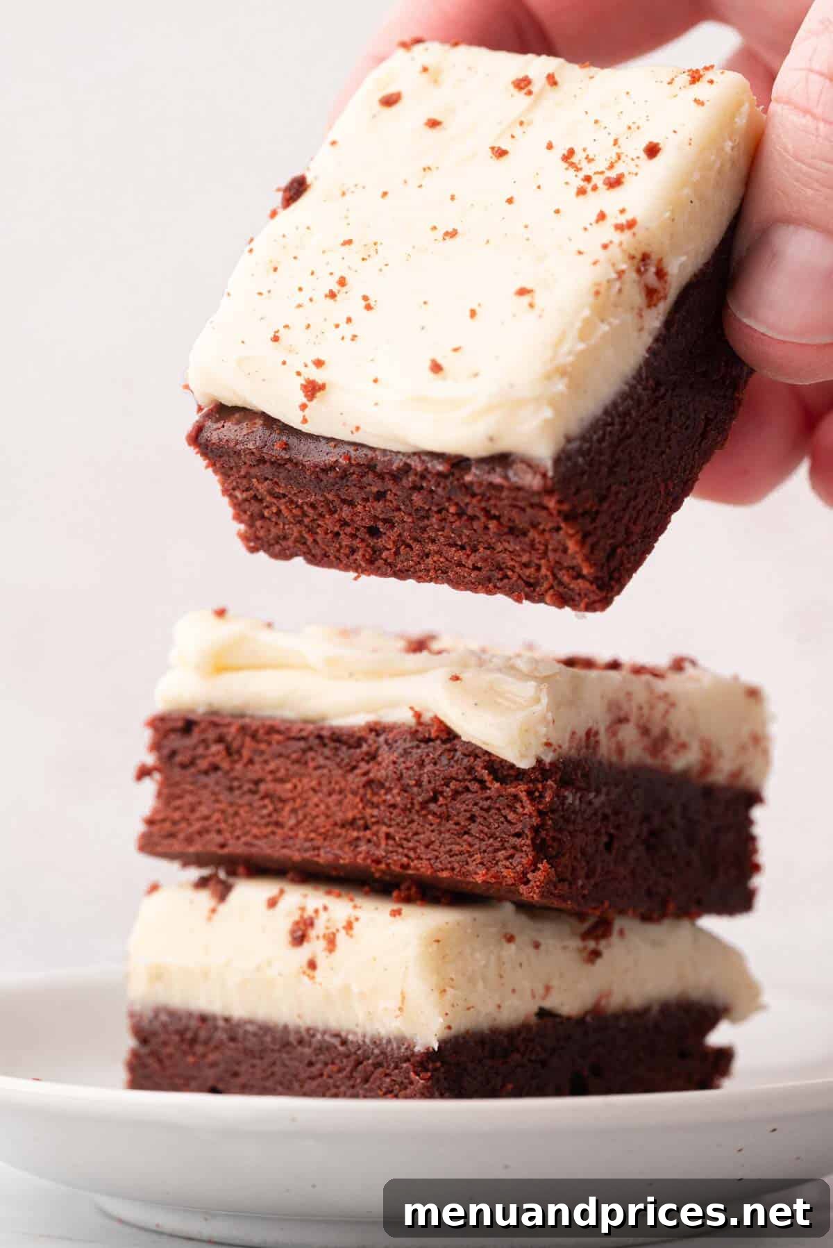Crimson Velvet Brownies 21 A hand lifting a red velvet brownie from a stack of three, showcasing the rich layers and creamy frosting.