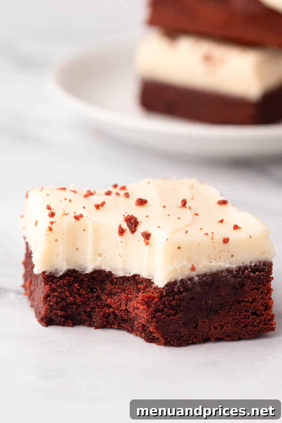 Crimson Velvet Brownies 22 A red velvet cake brownie with cream cheese frosting, showing a bite taken out, revealing its fudgy interior.