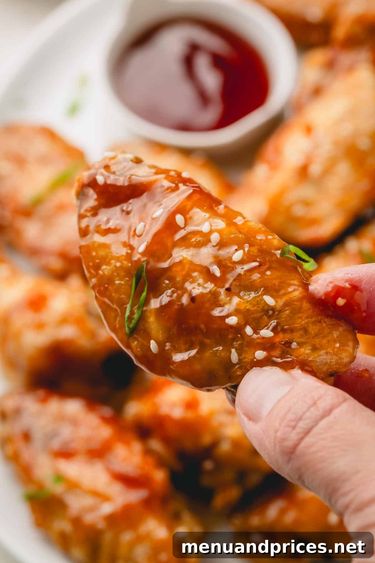 Sticky Teriyaki Chicken Wings 4 A hand holds a single, glistening honey teriyaki chicken wing, showcasing its crispy skin and sticky sauce, adorned with white sesame seeds and finely chopped scallions.