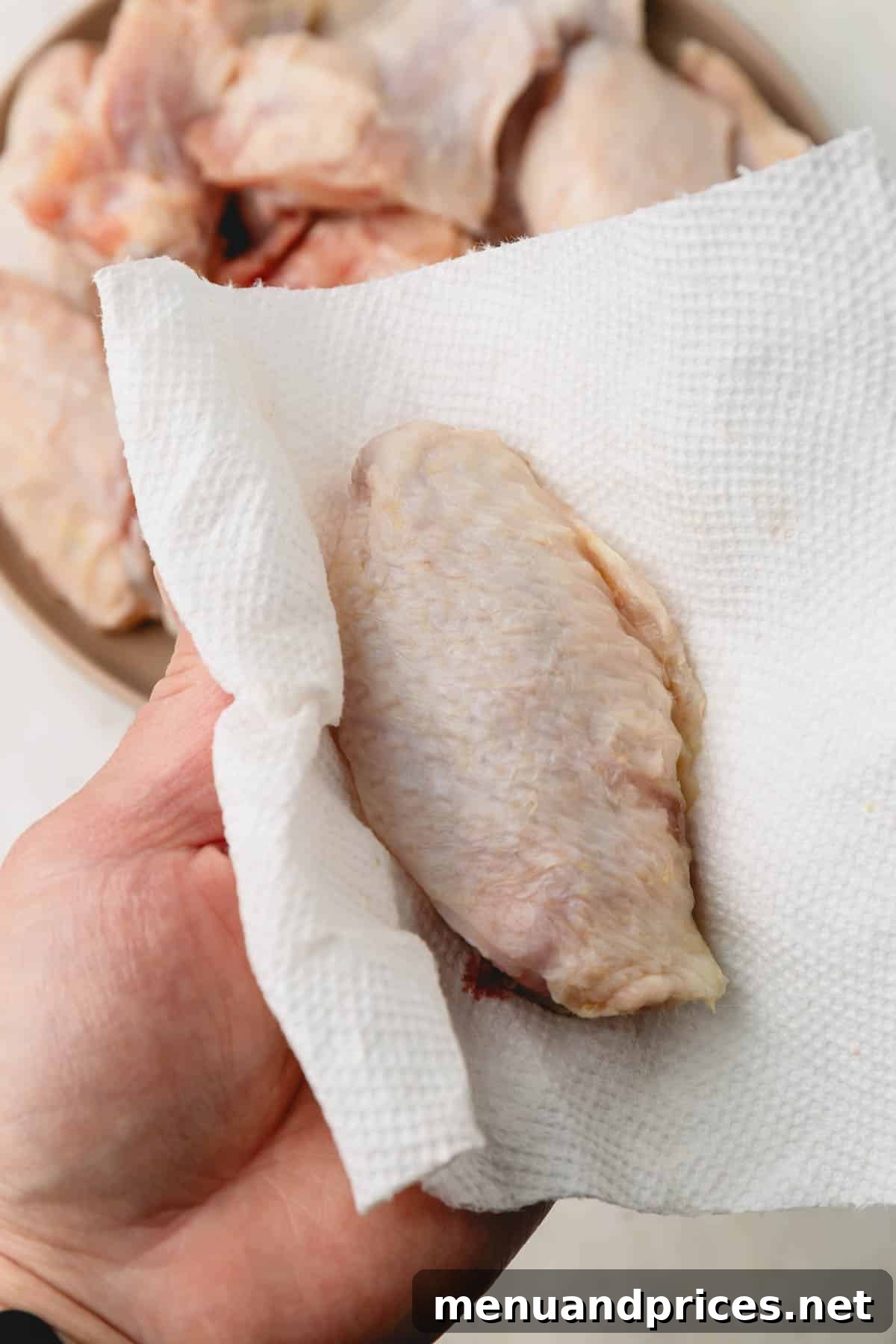 Sticky Teriyaki Chicken Wings 6 A close-up shot of a hand gently patting a raw chicken wing dry with a paper towel on a cutting board, emphasizing the importance of removing moisture before baking.