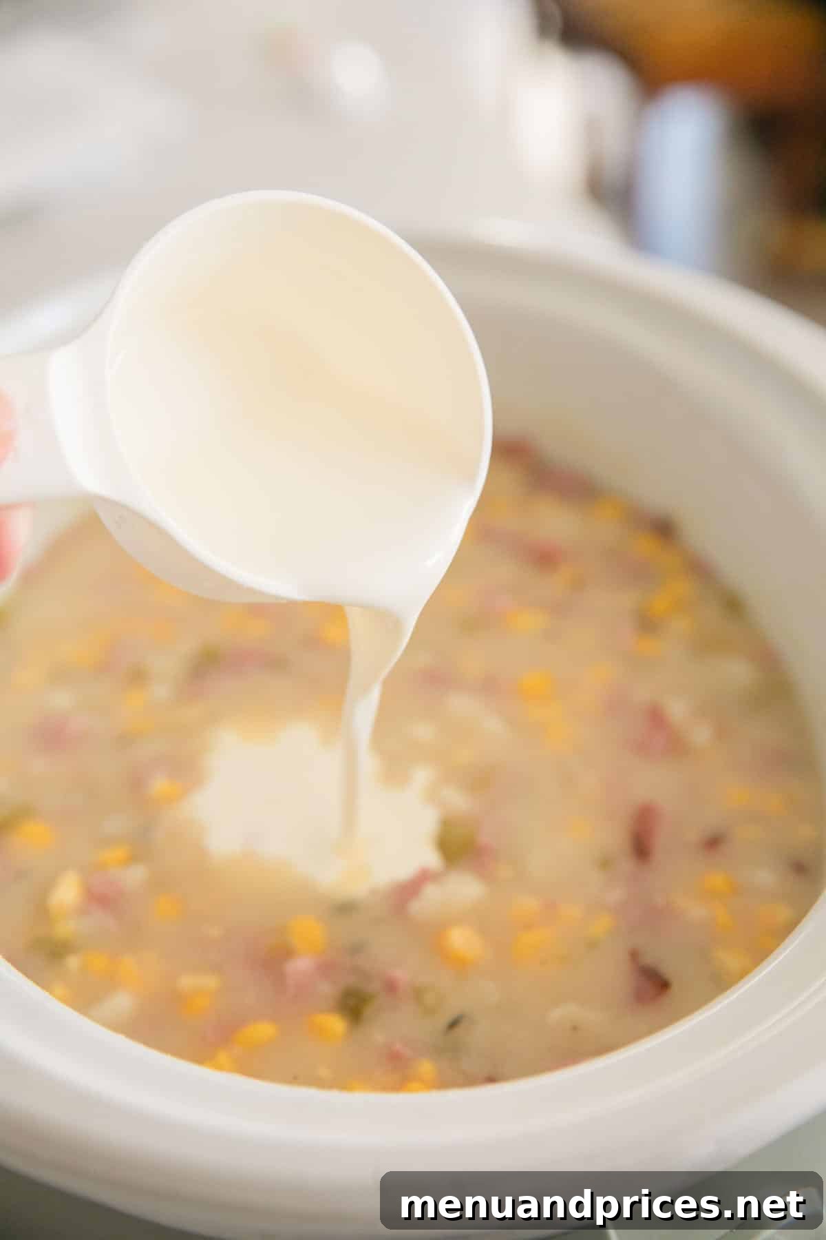 Hearty Slow Cooker Ham and Potato Chowder 9 Cream is being added using a white measuring cup into the slow cooker ham and potato soup.