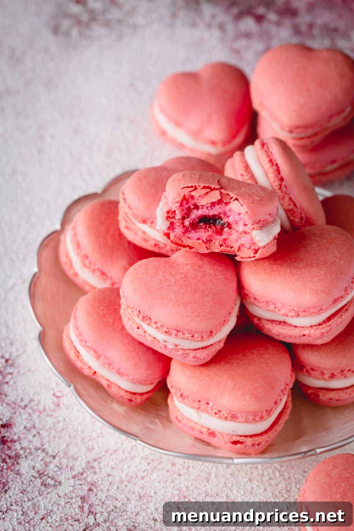 A stack of pink heart macarons with one macaron bitten into, revealing its jam and buttercream filling.