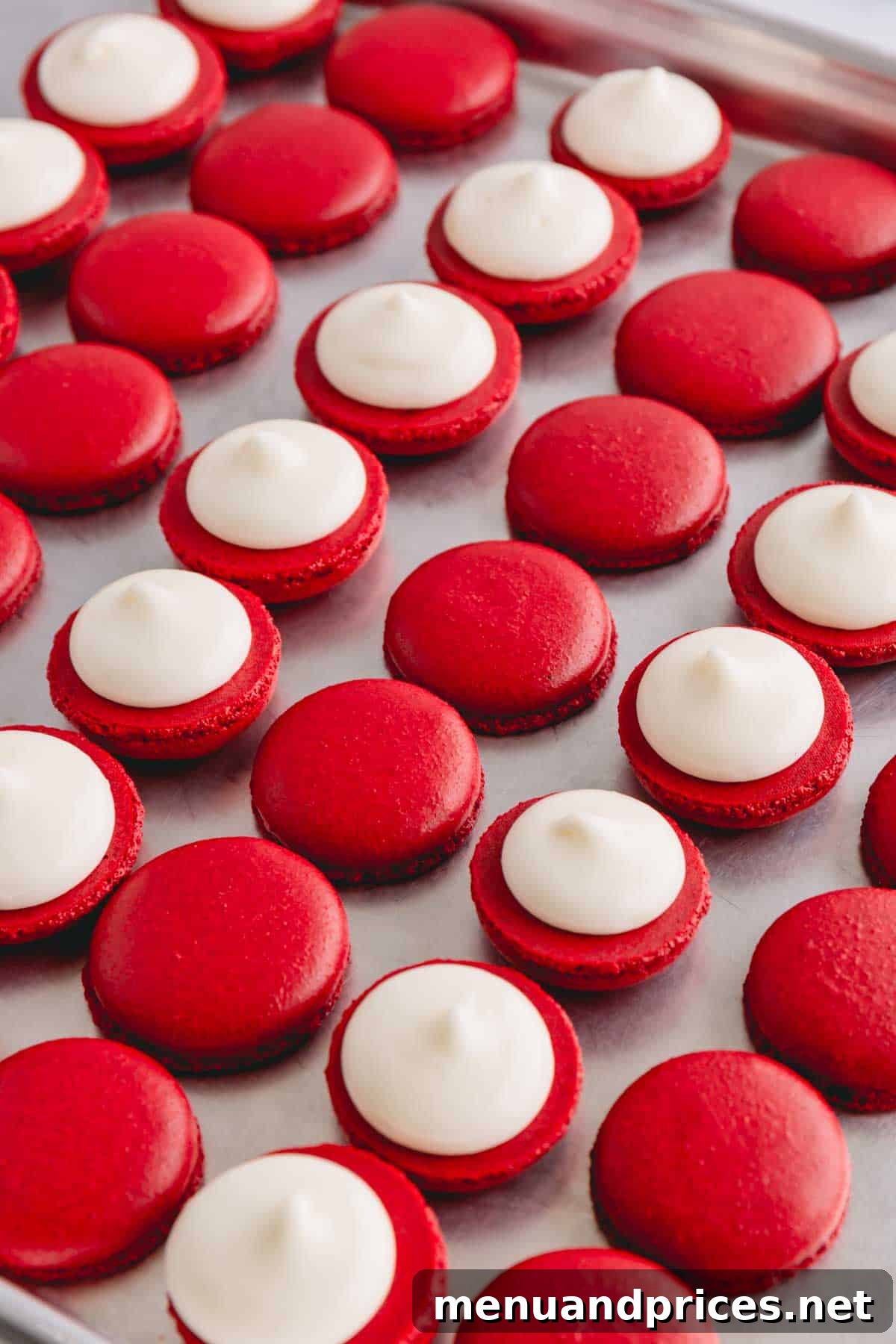 Red velvet macaron shells arranged on a baking sheet and half of the shells with cream cheese frosting, ready for assembly.
