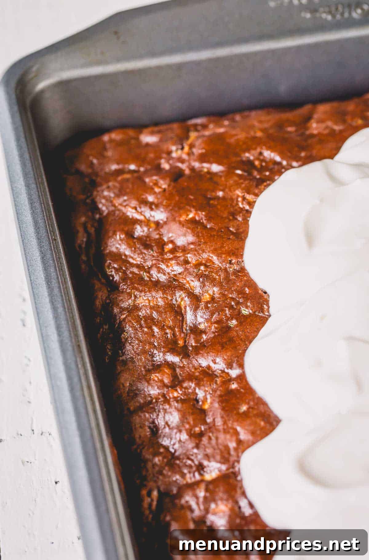 Fudgy Chocolate Zucchini Sheet Cake 3 Chocolate zucchini cake in a baking pan, with swirls of sour cream frosting being generously scooped on top.