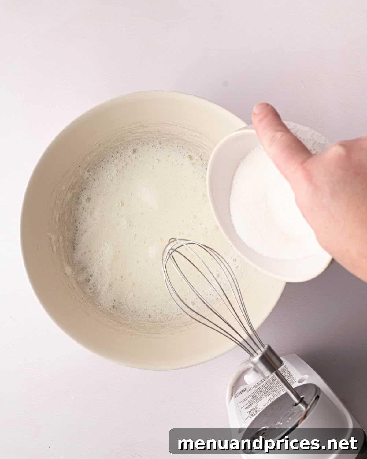 Zesty Lemon Dream 13 A white bowl with whipped egg whites and a hand adding a white bowl of sugar. A hand mixer is resting over the bowl.