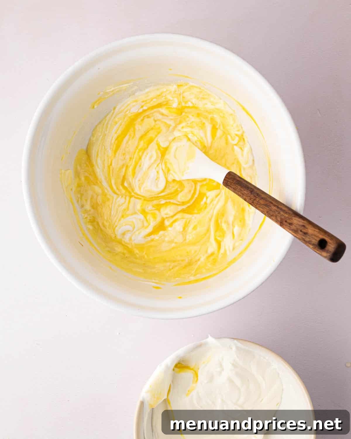 Zesty Lemon Dream 15 A white bowl with a white spatula and wooden handle resting on the side. The bowl has the thickened lemon base with whipped cream being folded in.