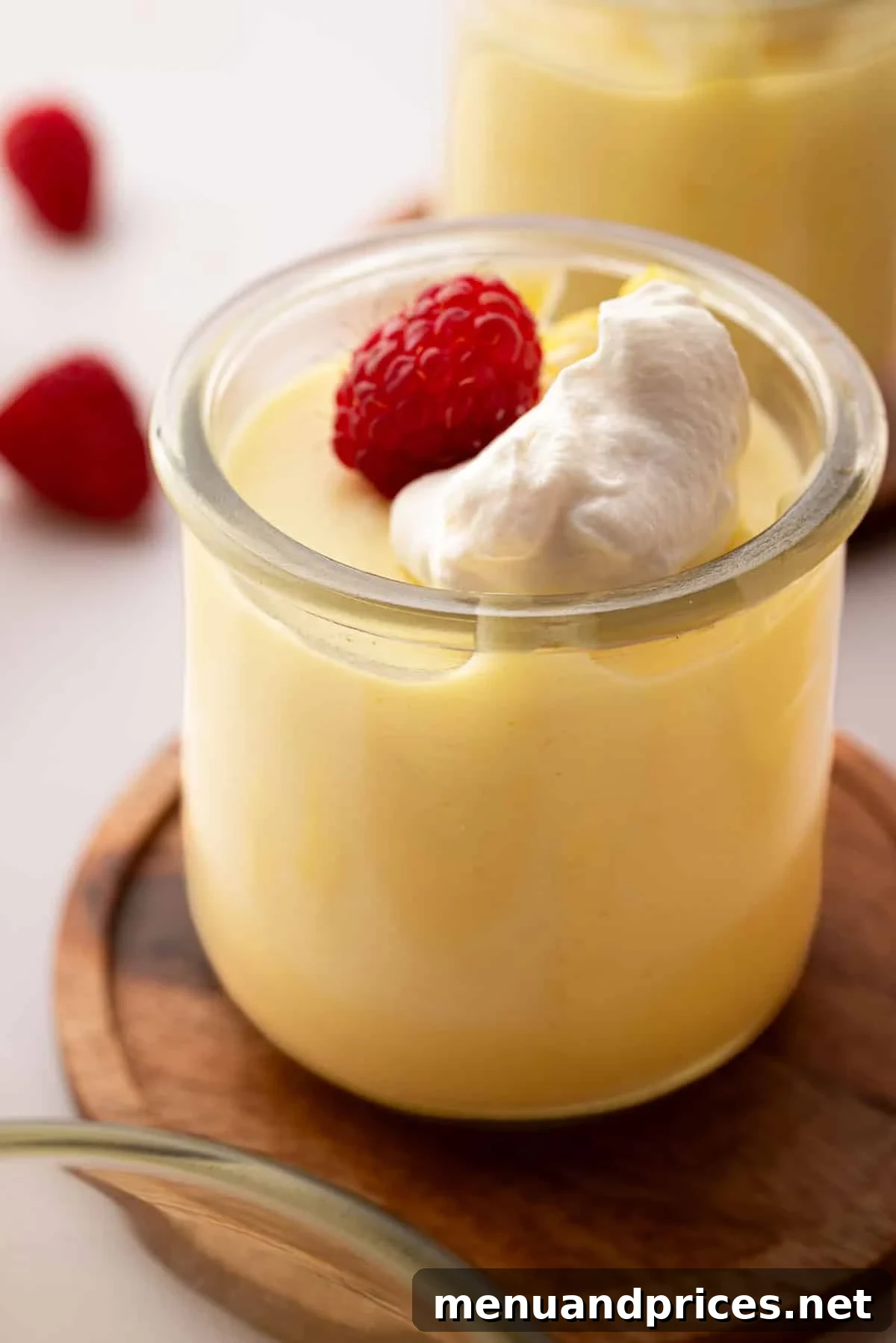 Zesty Lemon Dream 19 A small glass jar with lemon mousse and a raspberry on top. The glass jar is on a wooden coaster.